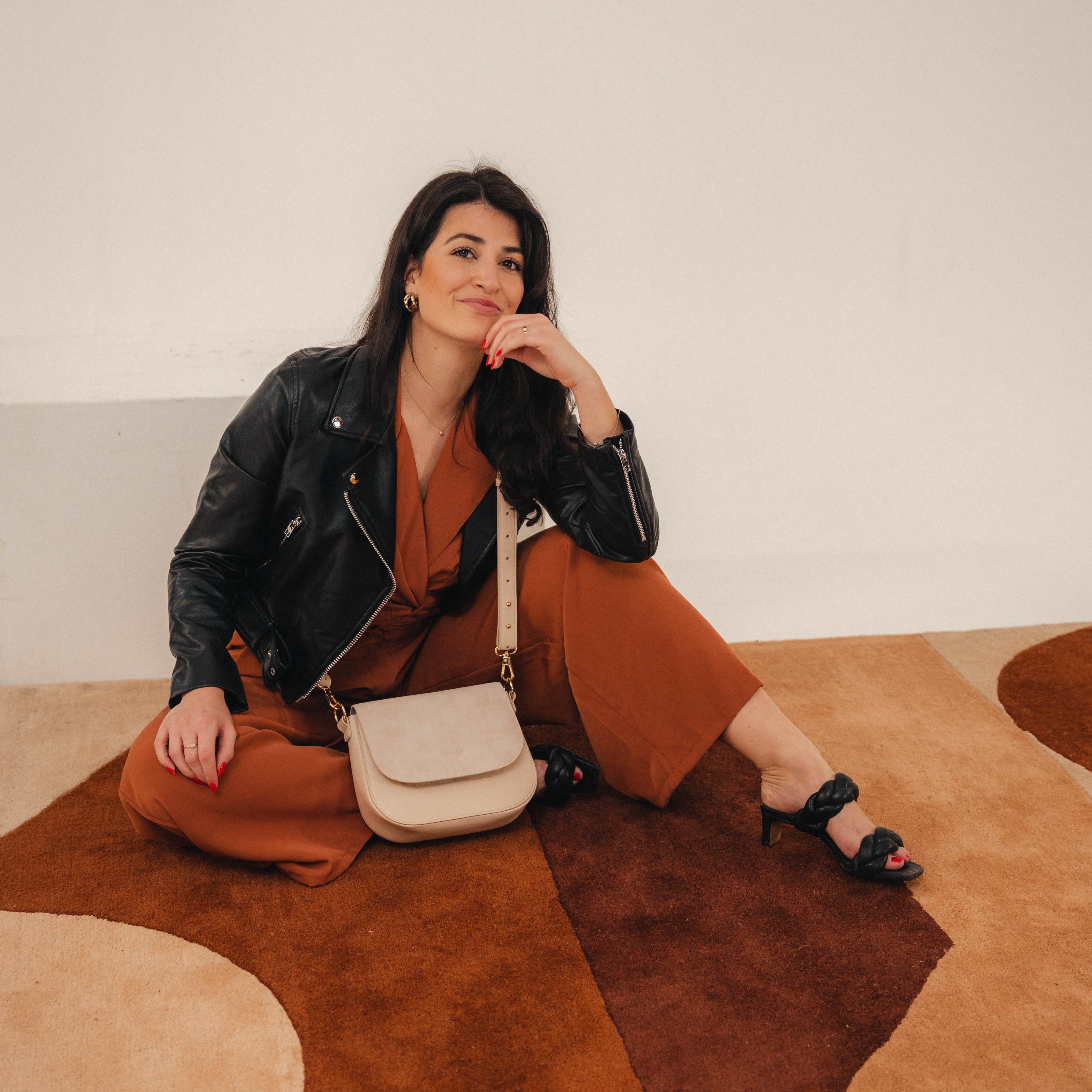 Frau mit schwarzer Lederjacke und rostfarbener Hose sitzend auf beigem und braunem Teppich mit beigefarbener Saddle Bag Louise Medium Umhängetasche