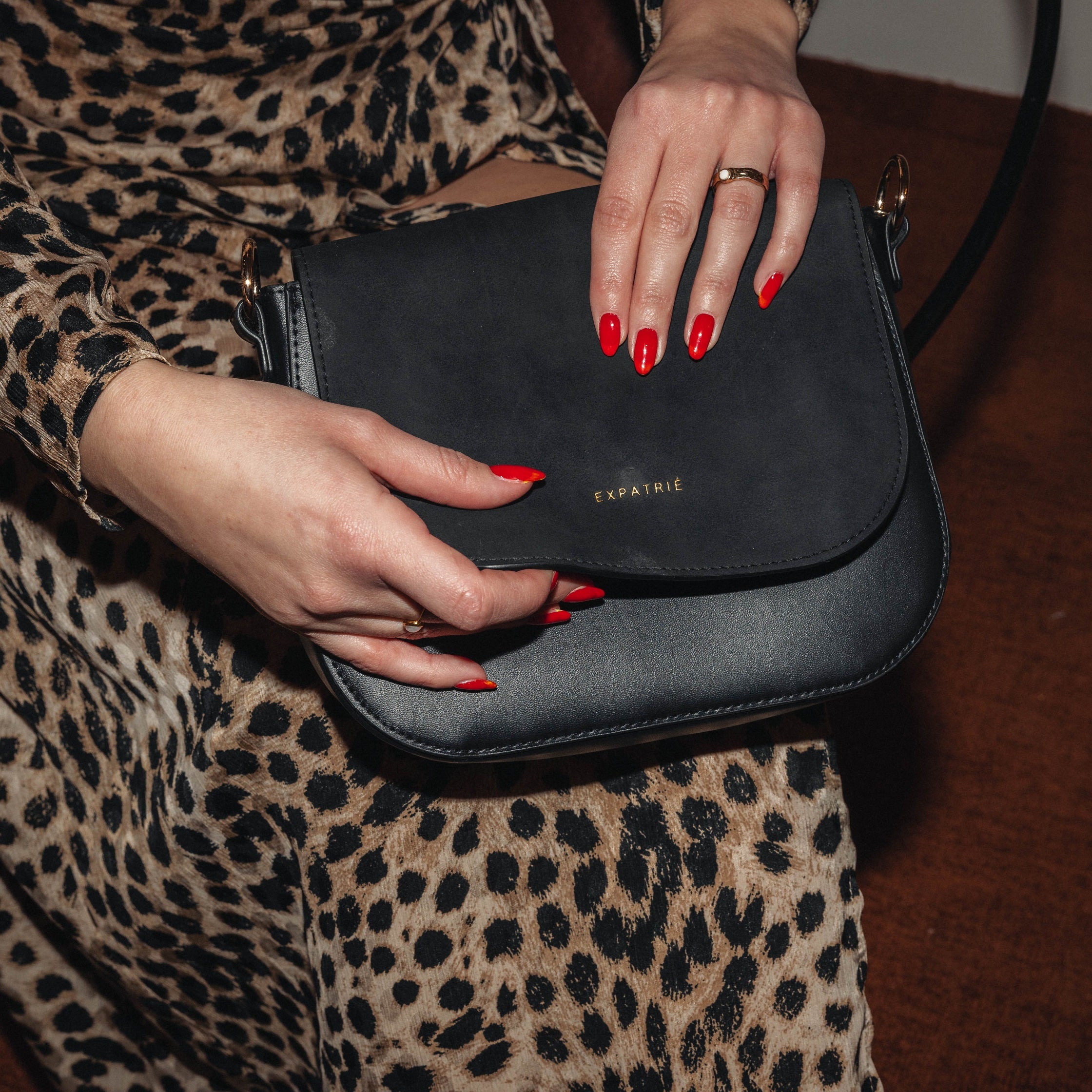Schwarze Saddle Bag Louise Medium von Expatrié aus Leder und Velours, gehalten von Frau mit Leopardenmuster-Kleid