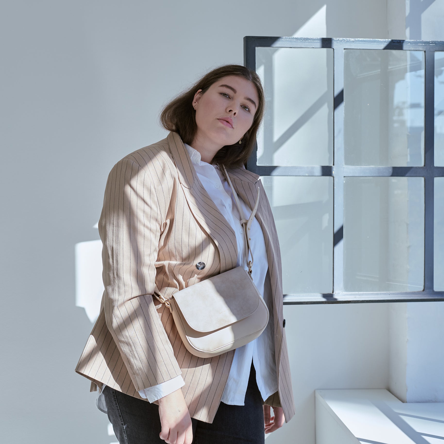 Frau trägt mittlere Saddle Bag Louise in Beige über Schulter, kombiniert mit gestreiftem beigem Blazer und weißer Bluse
