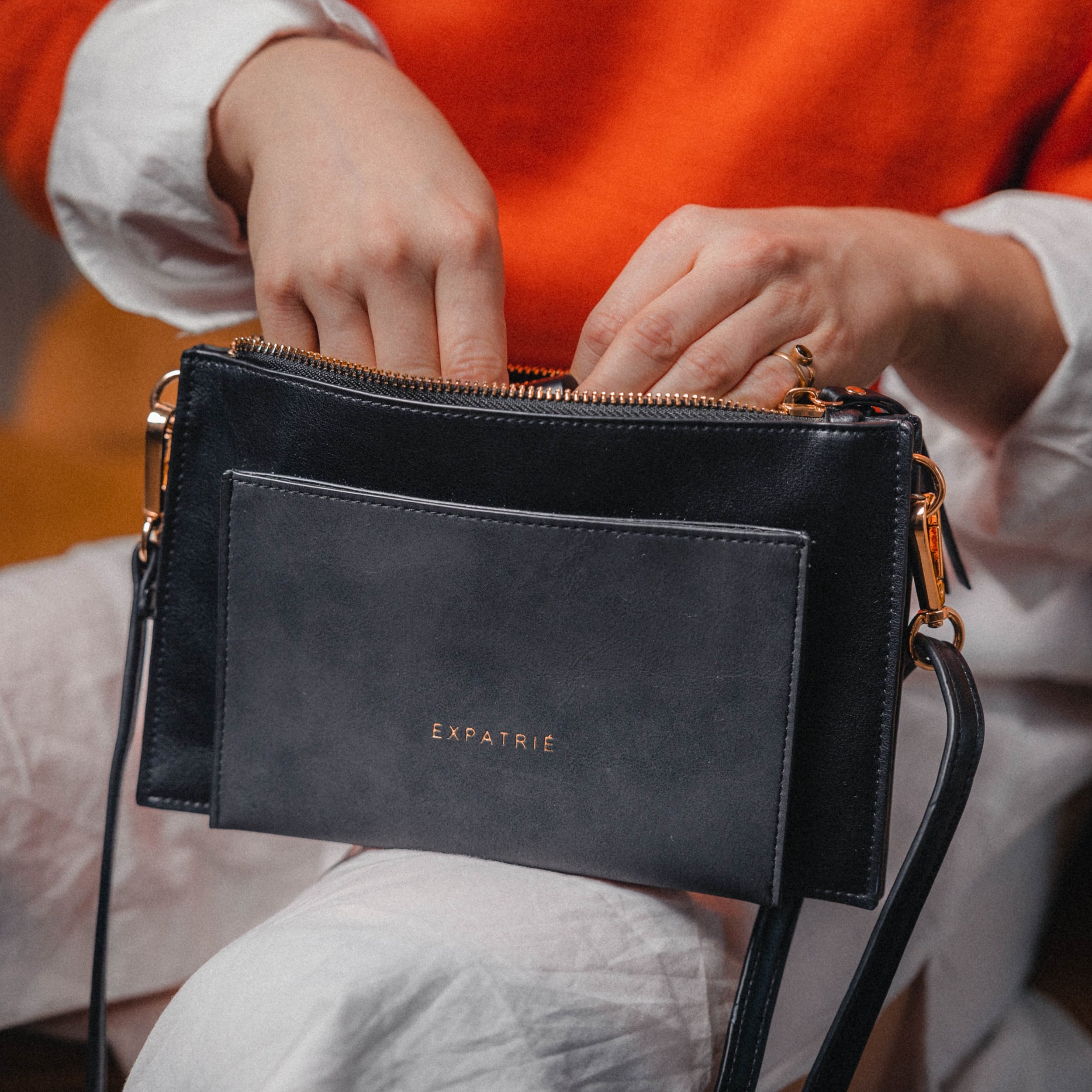 Schwarze Clutch 'Isabelle' von Expatrié aus Leder mit goldfarbenem Reißverschluss, getragen von einer Person in orangefarbenem Oberteil