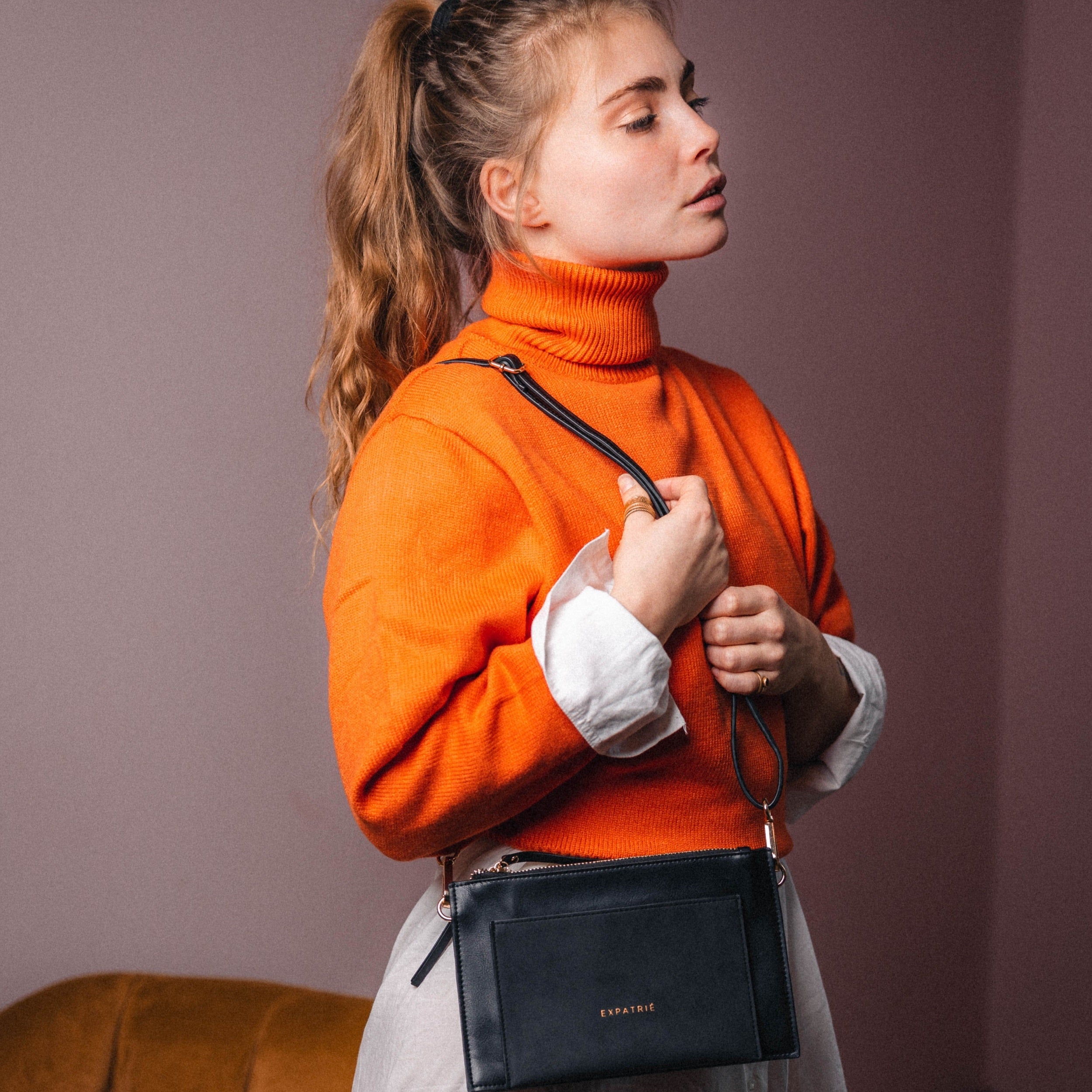 Frau mit orangefarbenem Rollkragenpullover hält schwarze Clutch Isabelle mit goldener Logo-Prägung