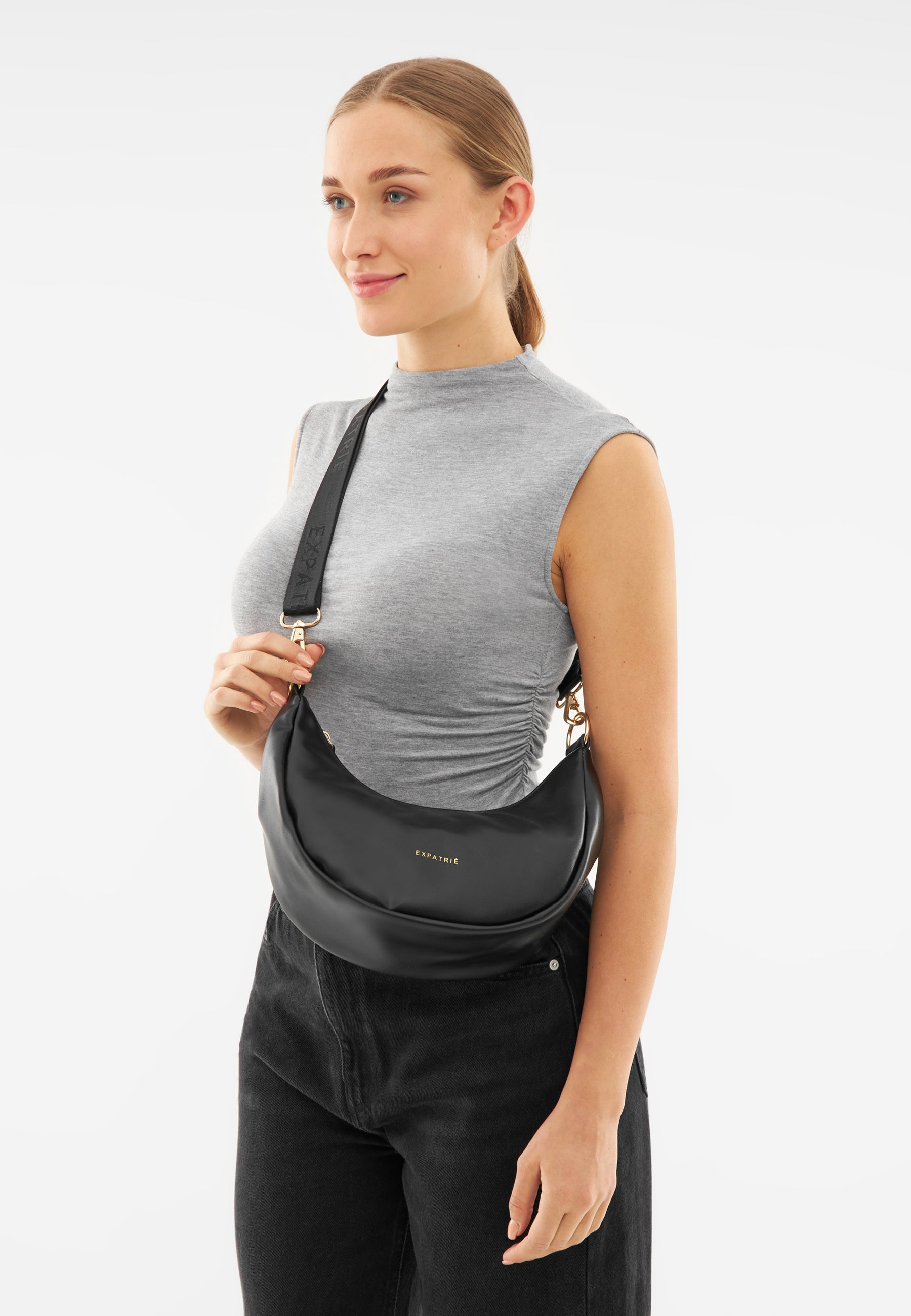 Frau trägt schwarze LEA SMALL Half Moon Bag von Expatrié mit breitem Schulterriemen und grauem ärmellosem Oberteil