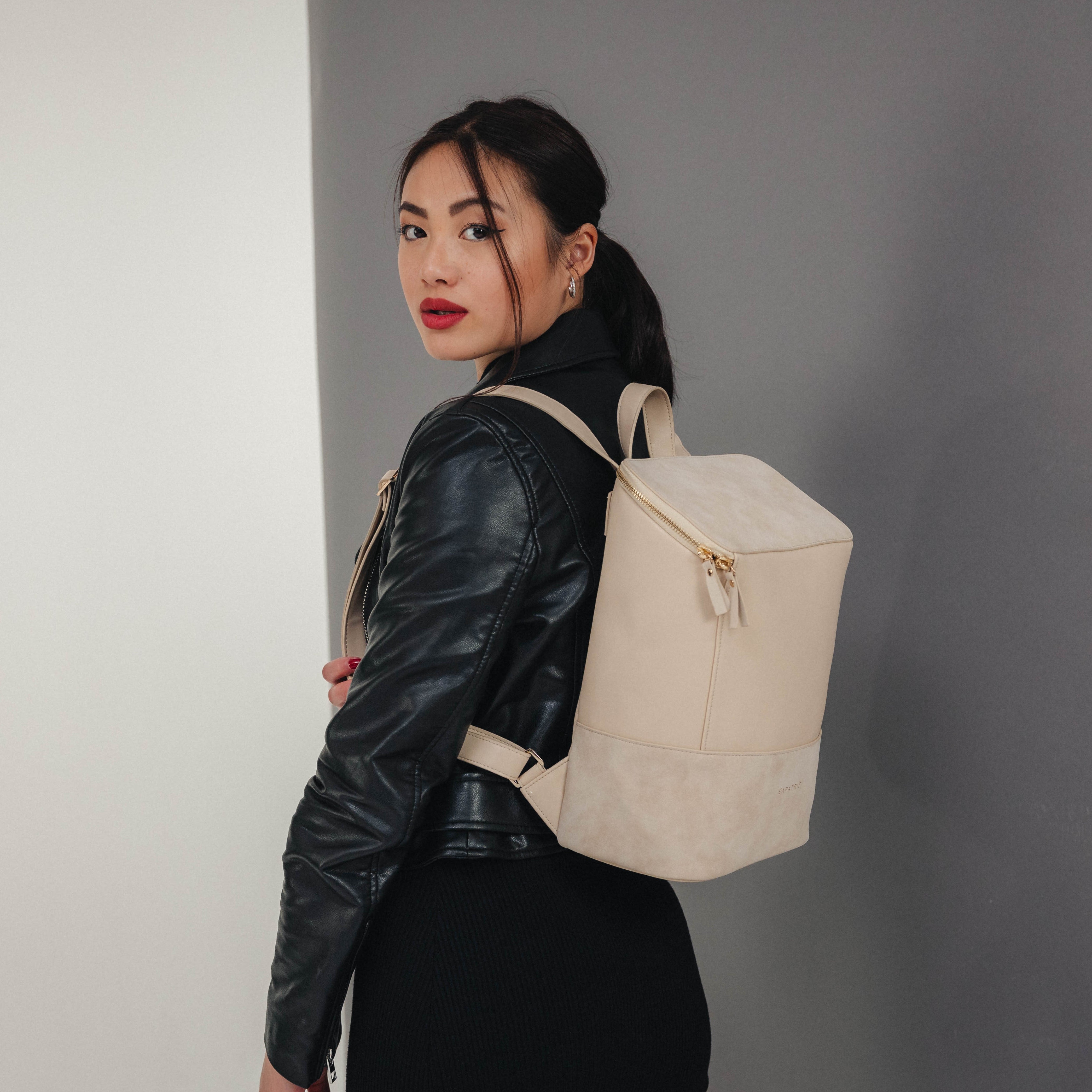 Frau mit Zopf trägt beige Lederrucksack Malou und schwarze Lederjacke vor grauem Hintergrund