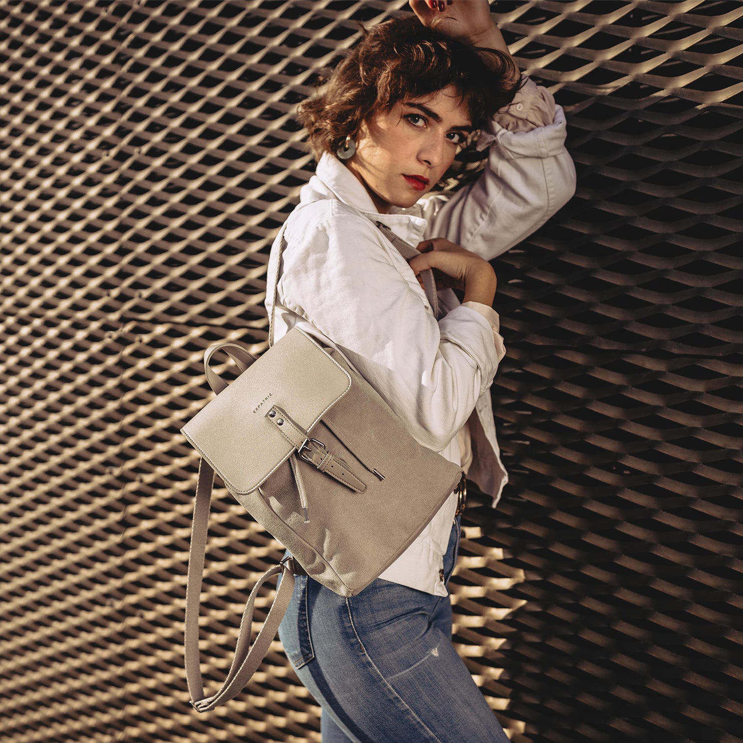 Frau mit hellem Mini Rucksack Anna in beige, weiße Jacke, blaue Jeans, vor strukturiertem dunklem Hintergrund