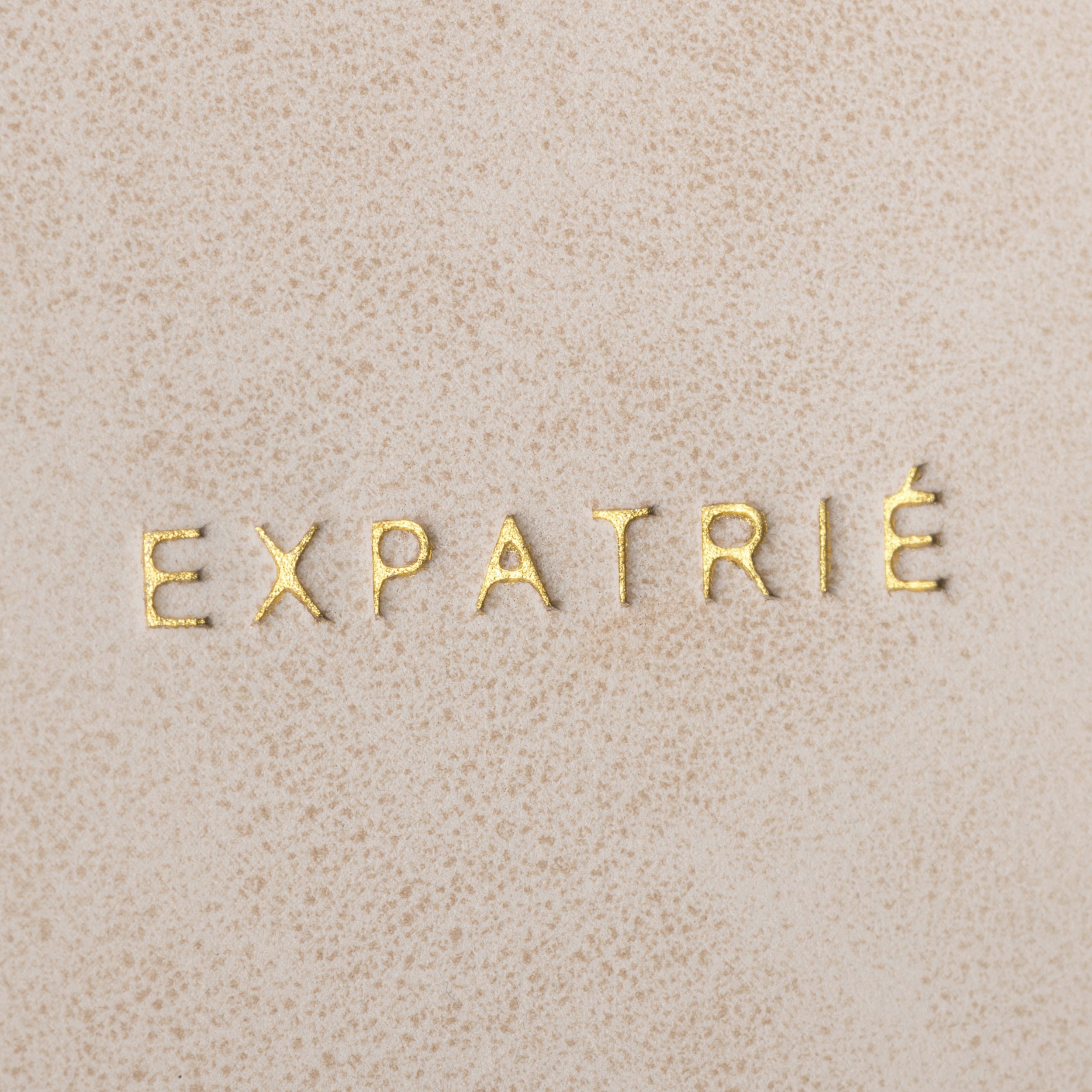 Goldene Buchstaben mit dem Text 'EXPATRIÉ' auf beigem Kunstleder des Rucksack Malou